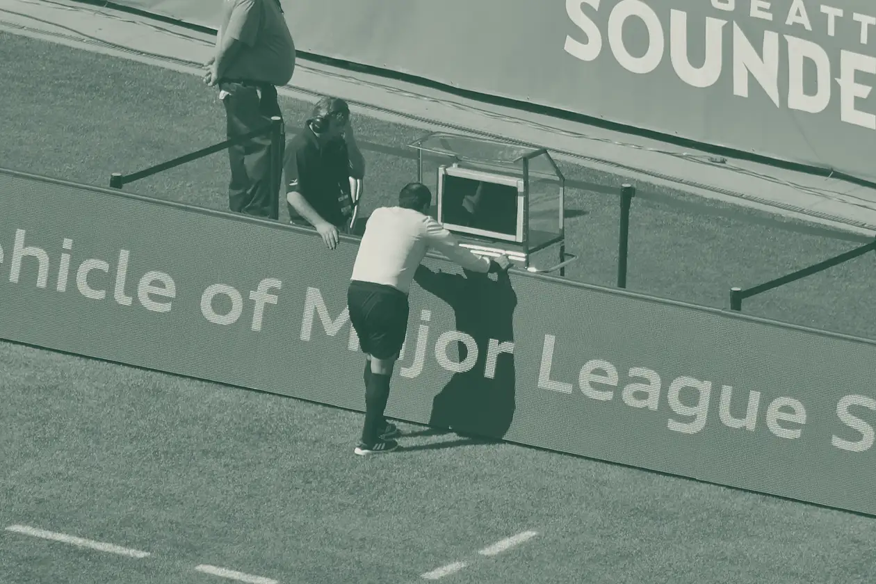 Árbitro de camisola amarela inclinado sobre o monitor do vídeo-árbitro junto à linha lateral, com um elemento da equipa de arbitragem ao lado