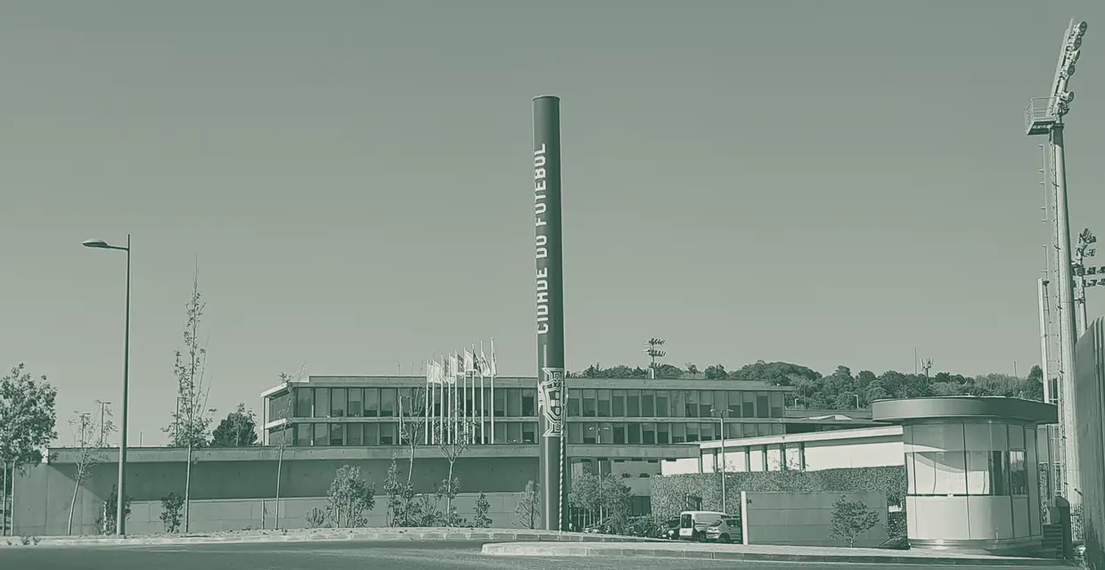 Mastro vertical com o nome Cidade do Futebol à frente do edifício da Federação Portuguesa de Futebol e de um campo com torres de iluminação